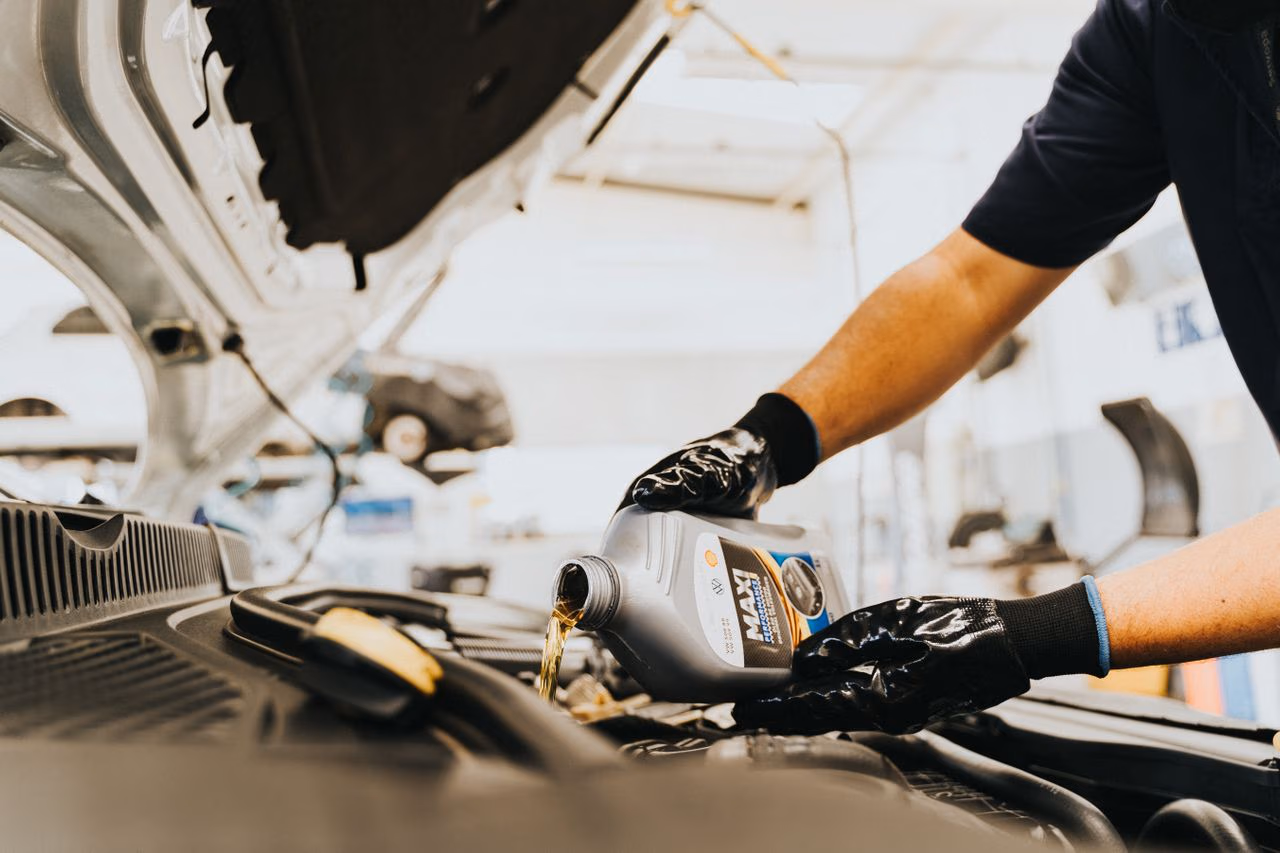 Person wearing gloves pouring motor oil into a car engine under an open hood.