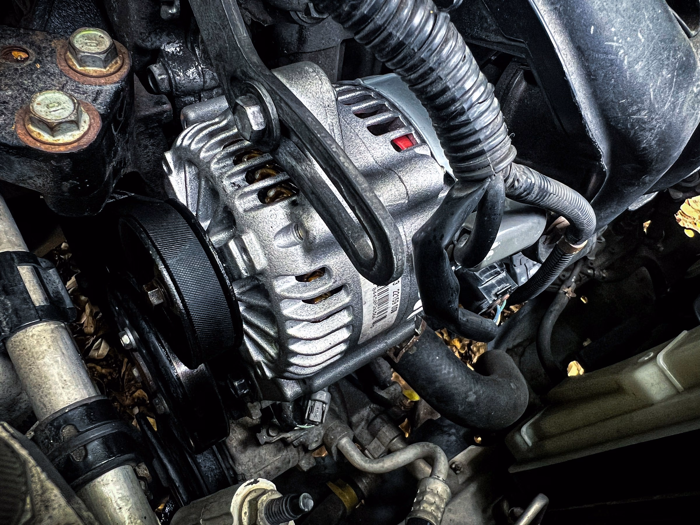 Close-up of a silver car alternator with attached black belt and surrounding engine components.