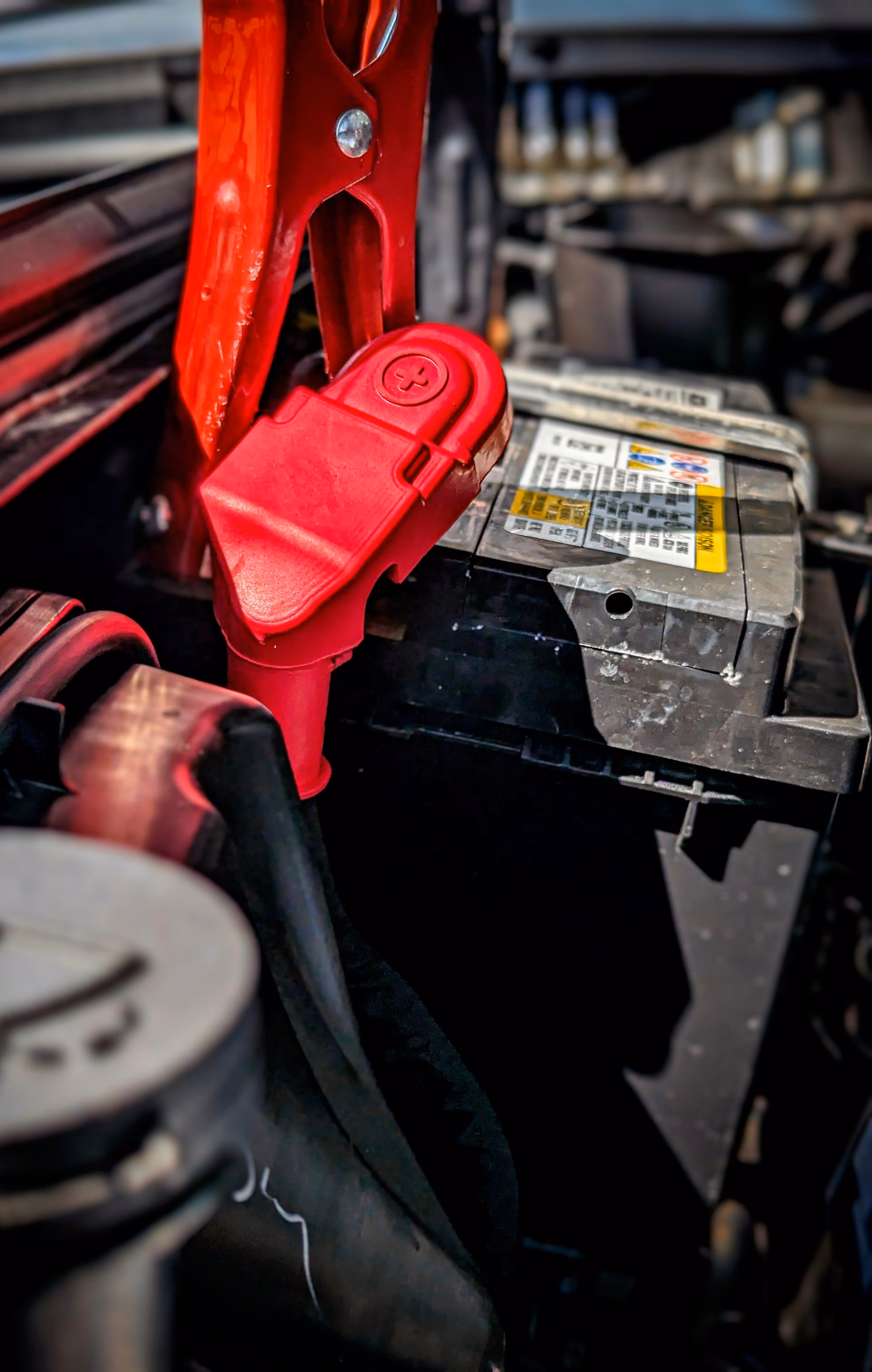 Close-up of a car battery with a red positive terminal clamp attached under a car hood.