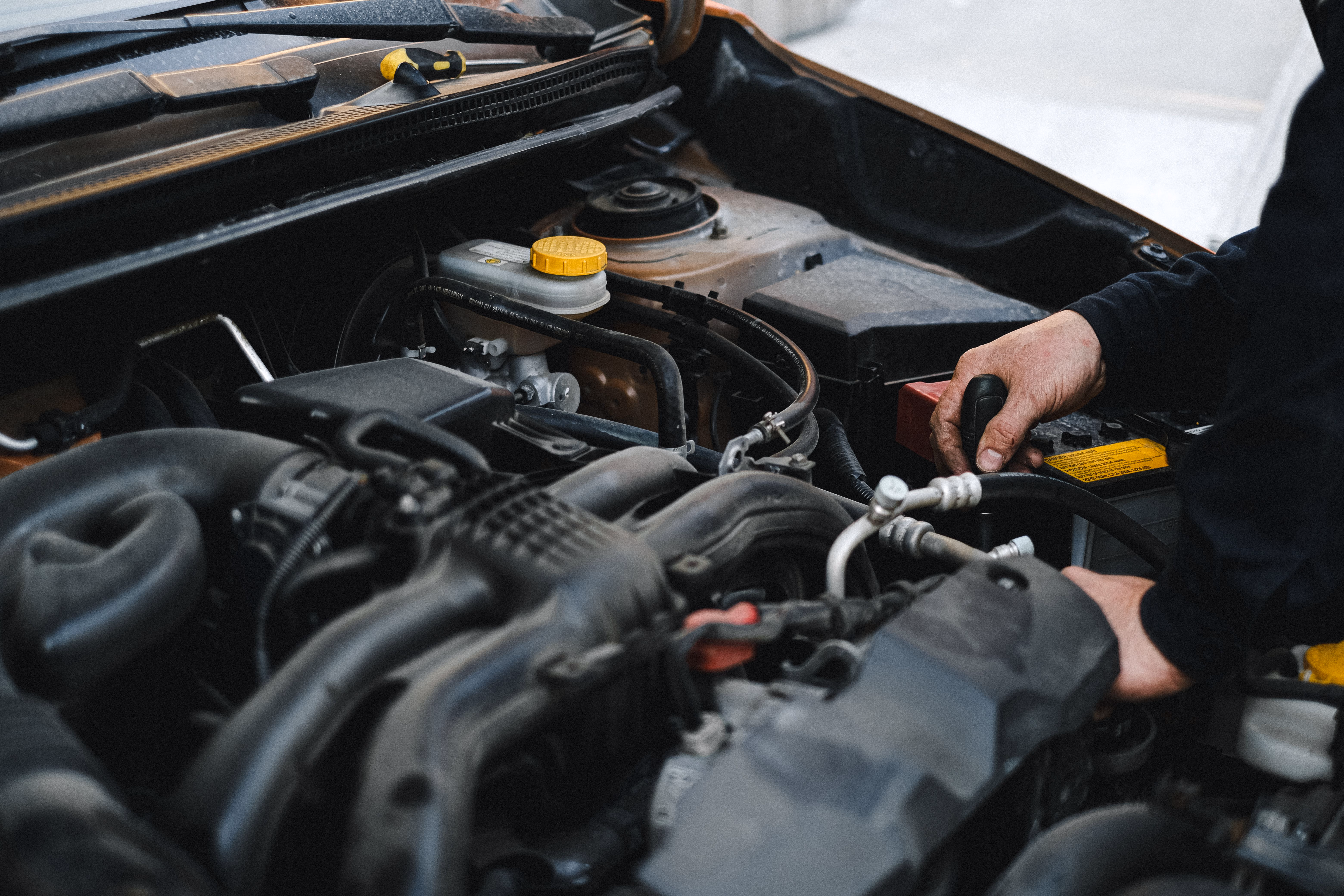 Person's hands working on a car engine with various hoses, battery, and fluid caps visible.