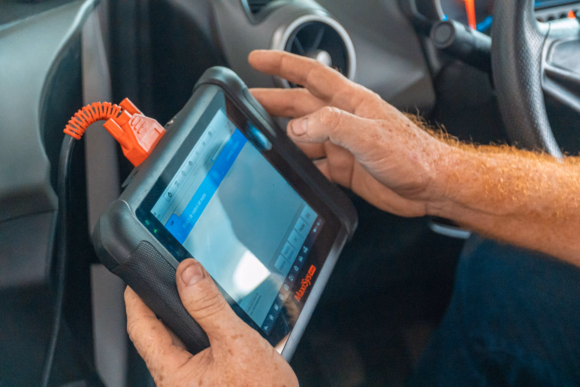 Person holding a MaxSys diagnostic tablet inside a vehicle, connected with an orange cable.