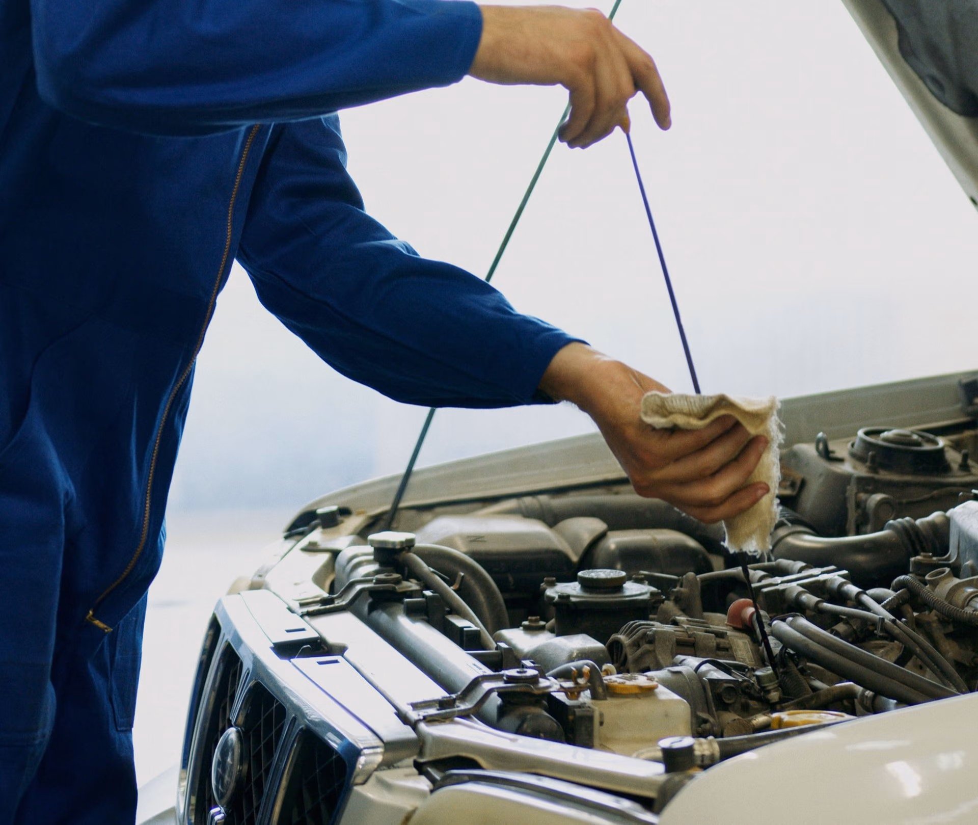 Mechanic in blue coveralls checking engine oil level with dipstick and cloth under car hood.