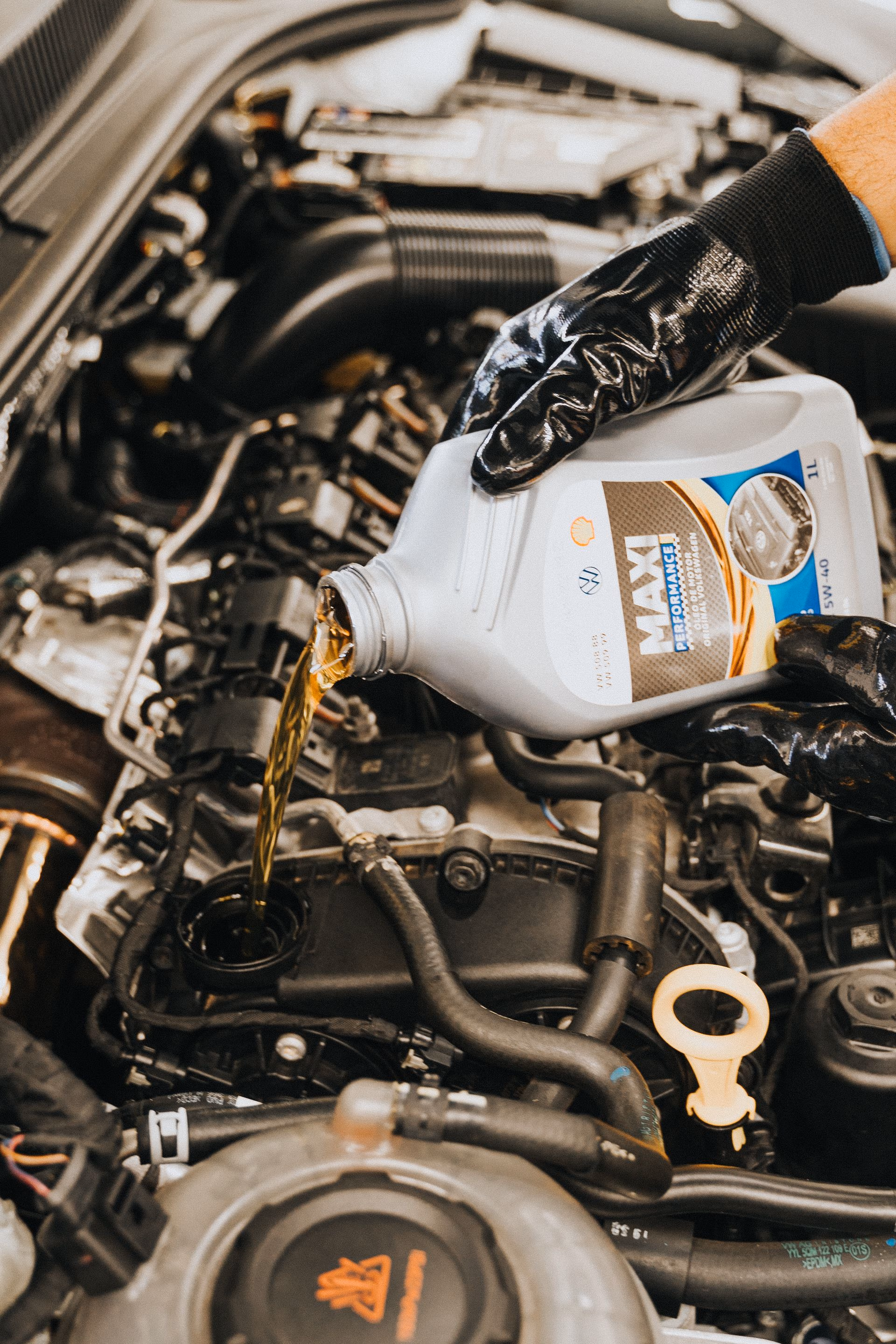 Gloved hands pouring motor oil from a Shell container into a car engine during an oil change.