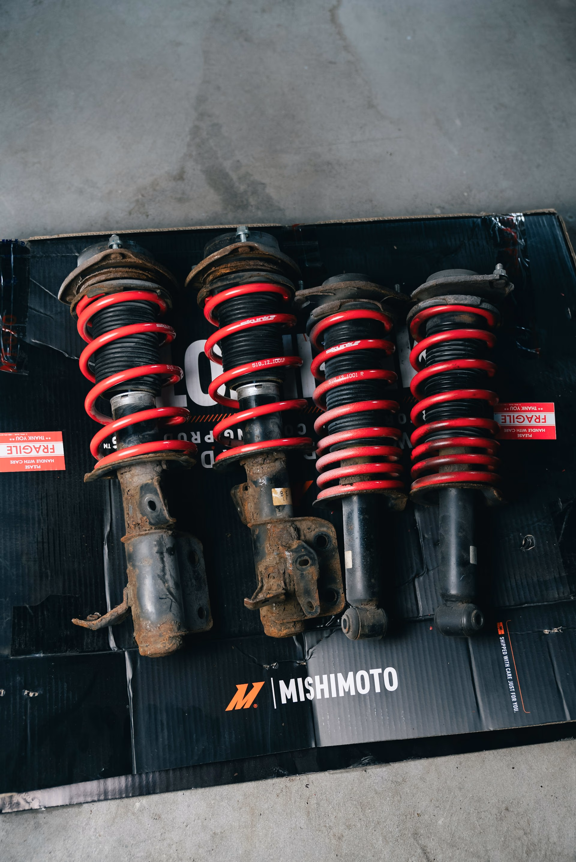 Four used car suspension struts with red coil springs on a black Mishimoto mat.