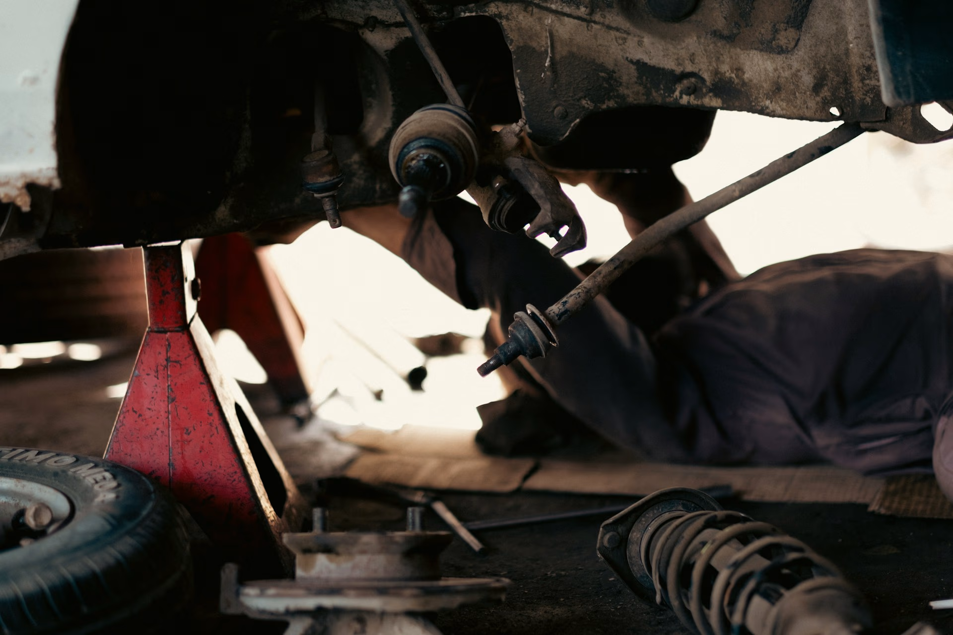 Mechanic working underneath a car on jack stands with suspension parts and a tire nearby.